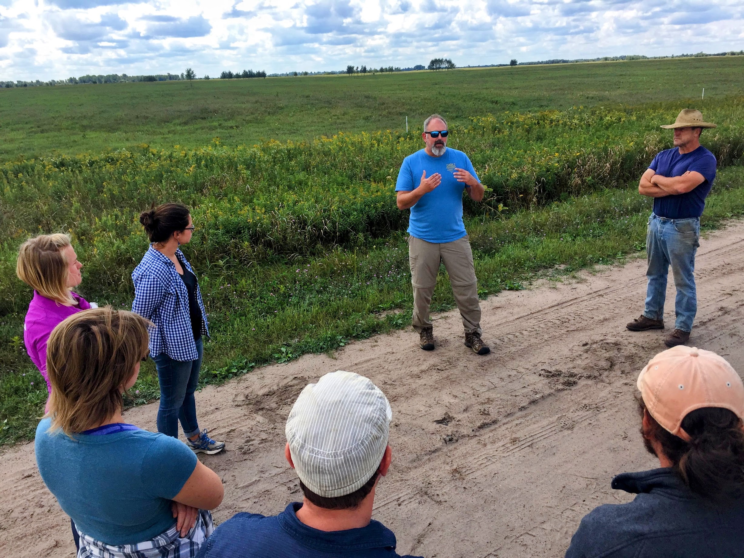 Randy Jackson speaks to a group of people on the edge of a field.