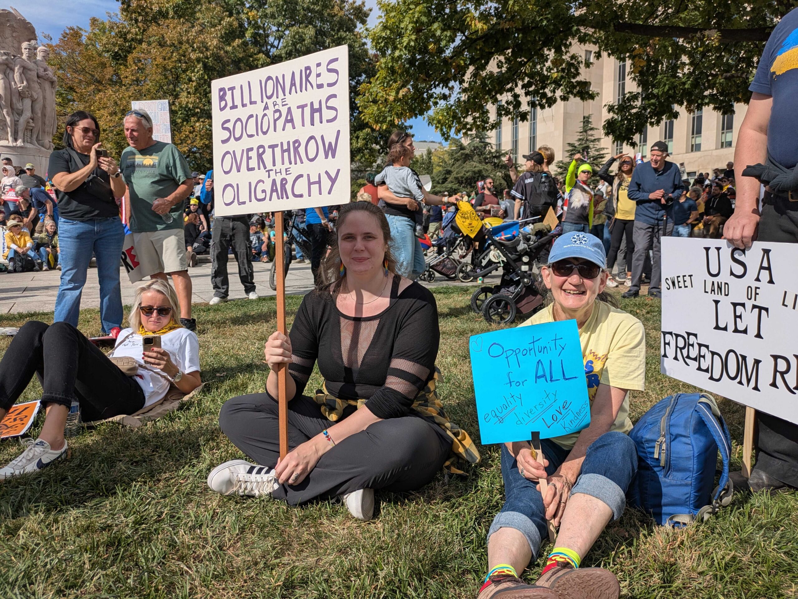 Emma Sutton, left, of Silver Spring, Maryland, sat in the grass along Pennsylvania Avenue holding a sign at the No Kings day rally in Washington, D.C., on Saturday, Oct. 18, 2025. (Photo by Ashley Murray/States Newsroom)