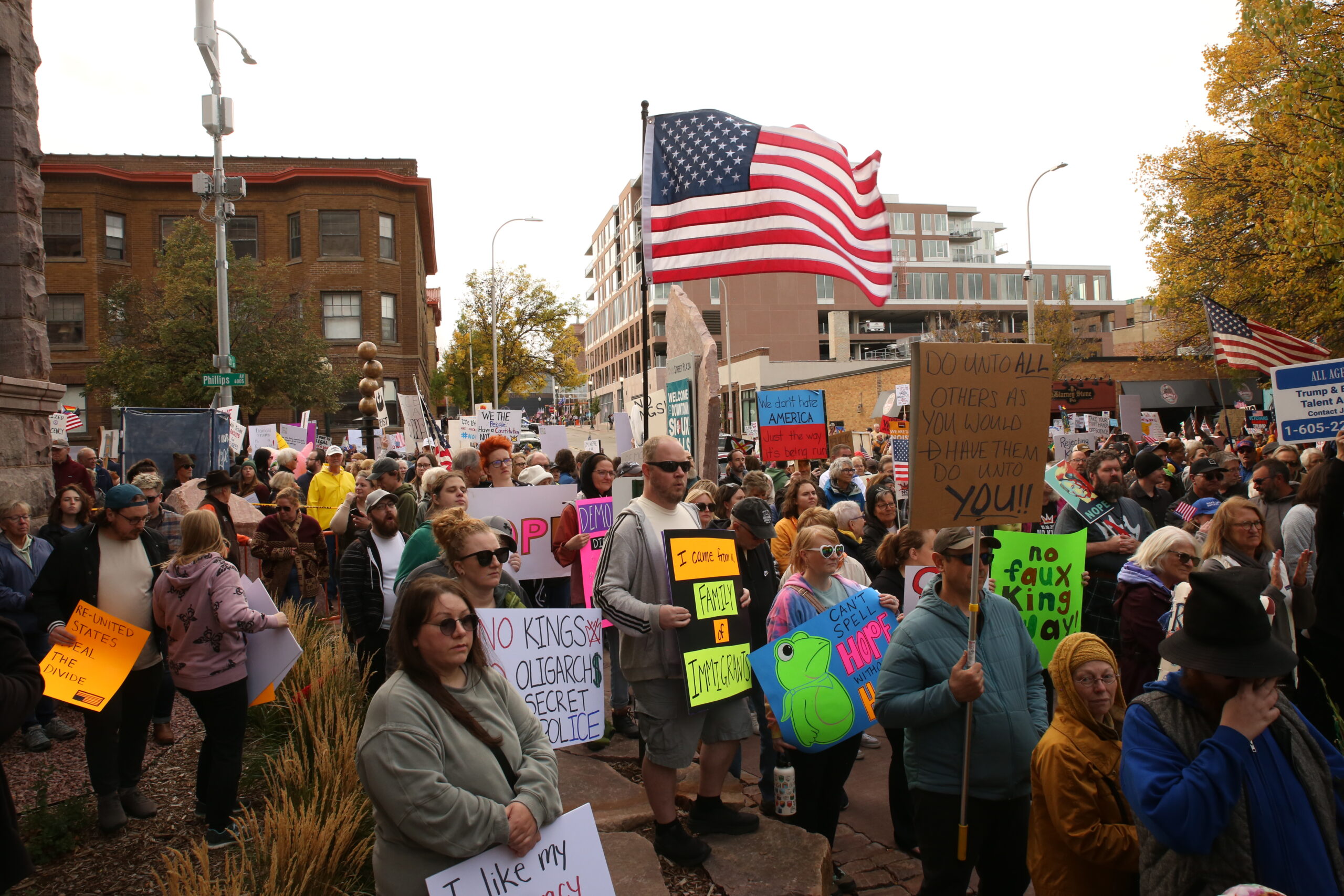 Protesters gather with signs and American flags while listening to speakers at the No Kings protest in downtown Sioux Falls on Oct. 18, 2025. (Photo by Makenzie Huber/South Dakota Searchlight)