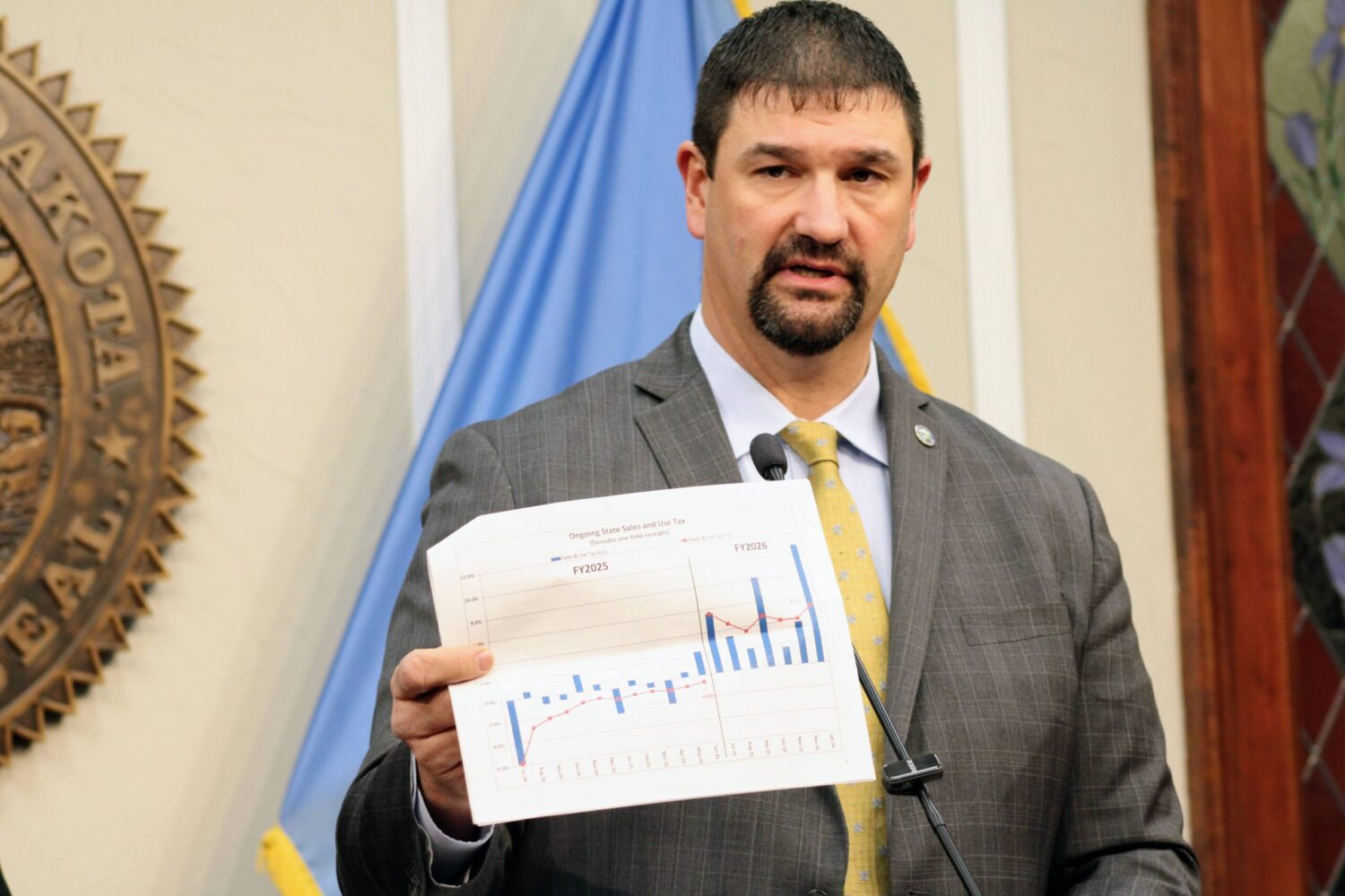 Jim Terwilliger, commissioner of the state Bureau of Finance and Management, holds up a sales tax bar chart while speaking to the media on Feb. 5, 2026, at the South Dakota Capitol in Pierre. (Photo by Seth Tupper/South Dakota Searchlight)